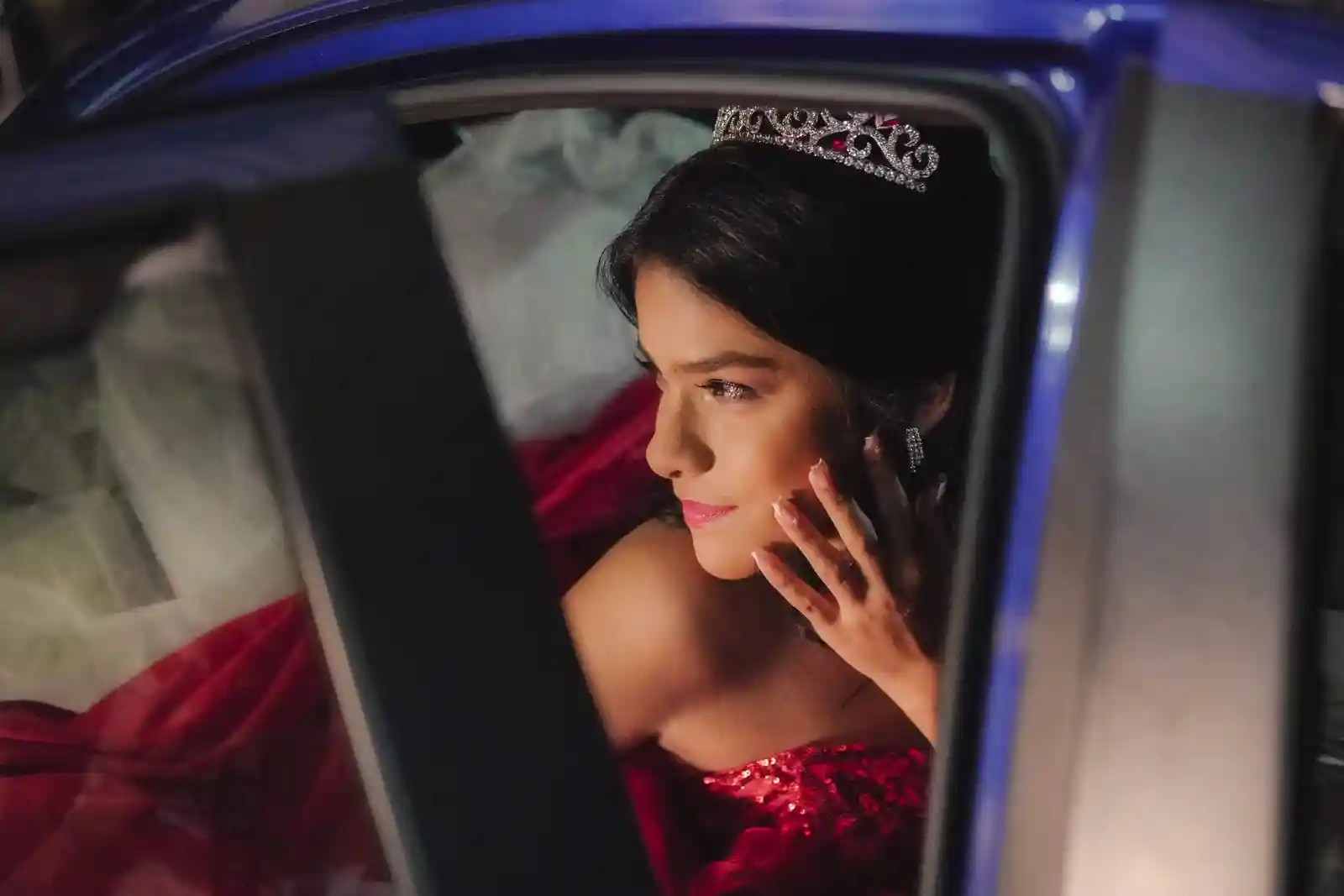 Quinceañera con tiara y vestido rojo, sentada en un carro azul, mira por la ventana.